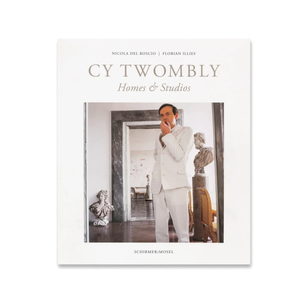 the cover of cy twombly homes and studios features a photo of the artist in a white suit standing with his hand at his moth, flanked by roman and renaissance busts in front of the marble doorway of his palazzo on via di monserrato, a painting by him barely legible in the room behind, from horst p. horst's iconic 1966 photoshoot for vogue