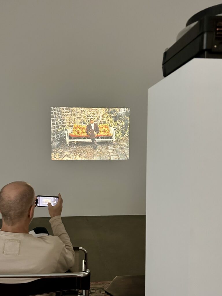 white guy in a wassily chair in david zwirner's la gallery taking a picture of a slideshow of william eggleston photos. the slide projected on the wall is of another white guy in a taupe suit sitting on an orange cushioned rocking sofa on a paved patio surrounded by a trellis, a 1970 photo taken of eggleston by a family friend using his camera, via wayne bremser