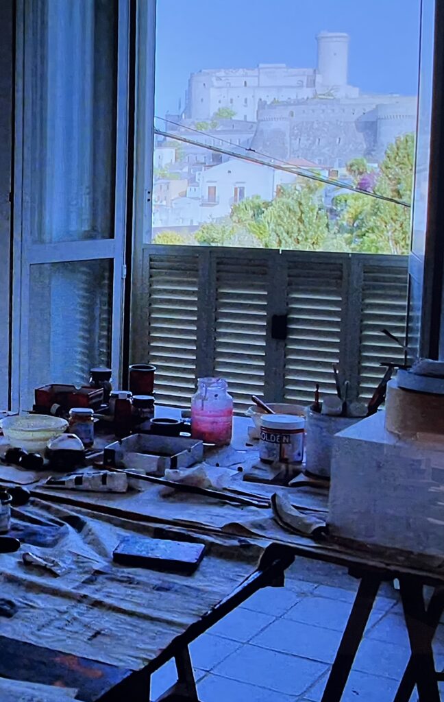 a screenshot of cy dear where the clutter of paint and brushes and an unfinished sculpture comprised of a little ziggurat of wood box, round biscuit tin, plate, and two spare rib-like plastery objects fill sawhorse tables in cy twombly's studio while the bay and fortress of gaeta are centered in the open window