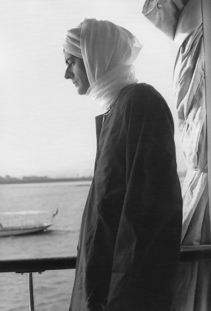 a  jan-feb 1962 photo by tatiana franchetti twombly of cy twombly wrapped around his head and neck like a sufi and wearing a dark caftan, standing in profile along the railing of the deck of their boat, with a furled canvas curtain behind him, and the water of the nile in the blurry background, on the shortest list possible for twombly's most epic fits