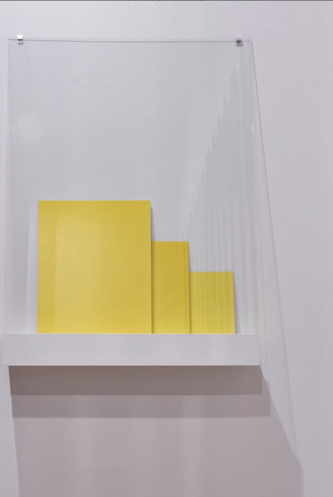 an installation of three liz deschenes monochrome yellow photographs, dye transfer prints with a viscerally powerful color in person, arranged so they slightly overlap each other as they lean on a white exhibition shelf, covered with a similarly leaning large sheet of glass pinned at the top where it meets the wall, at the george eastman museum in rochester, ny in january 2025