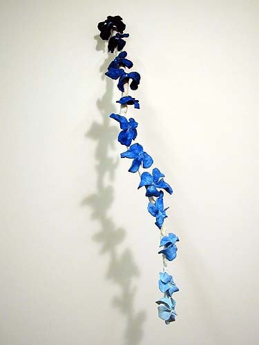 an arc of steel rod extends down and off a white wall. it is studded with sculpey flower blossoms, painted blue, gradating from dark at the top to light at the end. each flower was made within the run time of a disco song the artist erik hanson liked when he was a punk dj, so a playlist—and the listening to it—as sculpture.