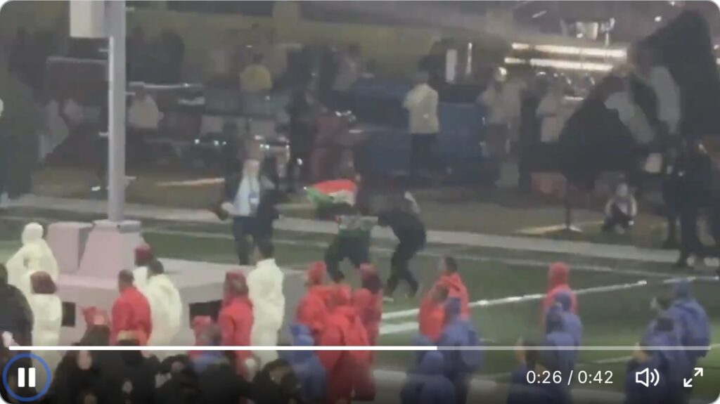 screenshot of a video from the superbowl halftime show depicting two agent smith type security agents in dark suits grabbing a dancer holding a palestine/sudan flag, about to tackle him to the ground on the edge of the football field. in the foreground are a couple dozen more dancers in formation, wearing red white and blue sweatsuits, waiting for their cue to start stepping. another dancer with a large black flag depicting the white silhouette of two little children reaching toward a capital a, a sign for A Minor, from kendrick lamar's not like us, posted to bluesky by jphillll