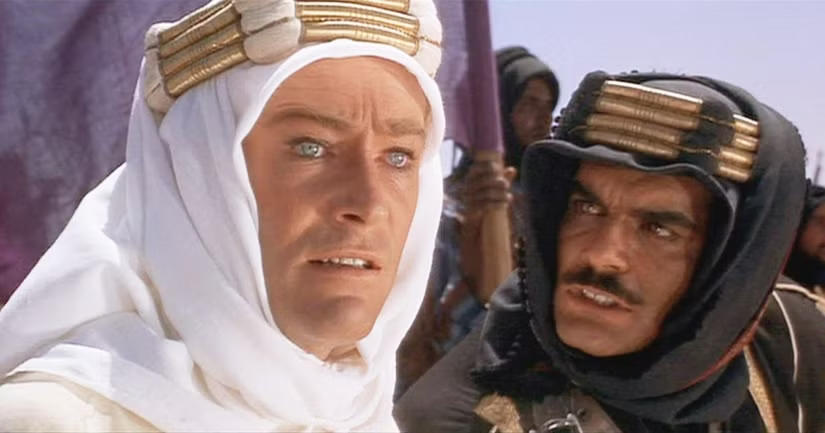 a promotional still from david lean's lawrence of arabia, released in december 1962, in which peter o'toole, staring intensely, lips apart, off to the right, wearing a white kaffiyeh and gold 'iqal, while omar sharif on his left stares intensely at him, lips apart, and wearing a black kaffiyeh and gold 'iqal