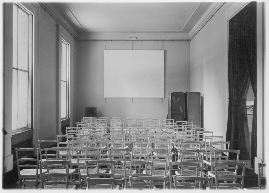 a black and white photo from 1938 of a tall ceilinged gallery in the musee du jeu de paume in paris set up as a screening room. thick dark curtains on the doorway on the right wall are currently open. 75 or so simple wooden cafe chairs are arranged in twelve rows facing the far wall, on which a white screen that looks a lot like a robert rauschenberg white painting, is hung high on the wall. the dimensions are for 16m, 4:3, basically. a folding screen stands in the corner below it. on the left wall, two giant windows let in all kinds of beautiful daylight, which, I must admit, is not something I'd have considered for a screening room. via moma archive