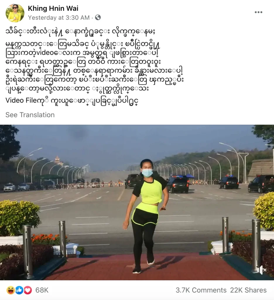 screenshot of burmese fitness instructor khing hnin wai's facebook post from feb 1, 2020 with burmese text over a video still of khing doing an exercise routine in a traffic island as a stream of black military and police vehicles drive behind her on a wide, blocked road leading to the national legislature. 