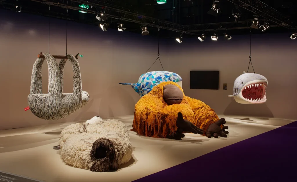 a low platform and a mushroom colored corner with a deactivated flatscreen at the design miami/basel fair in 2018 are the setting of this press photo of five giant, shaggy and/or plush furniture objects in the shape of endangered species, including three hanging: a sloth, a blue whale, a great white shark; and two flop lounging: an orangutan that wouldn't fit in a sprinter van, and a polar bear on its back in a nsfw way. the objects were made by a white south african artist who goes by porky hefer, which may be his name, and it was organized by some art advisory people, and sponsored by the leo dicaprio foundation as some sort of greenwashing thing, where some of the proceeds of sales of these monstrosities went to leo's ngo, but what happens if they don't sell? these ed. 1s sit in storage for seven years and then turn up at a random auction. which happened to the shark as i type this in feb 2025. image by james harris via wallpaper magazine, btw, what a commendation of journalistic integrity. but at least pr photos don't get deleted after the license expires in a year, so thanks for that