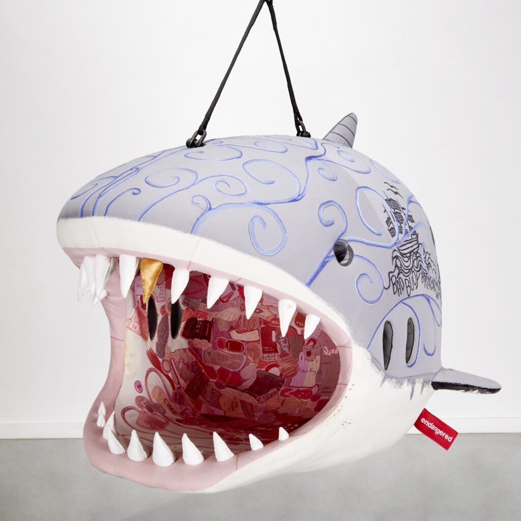 a plush shark embroidered with graffiti and save the earth type messages hangs from a two-point harness, its mouth wide open because that is where you're supposed to sit. it is a hanging chair, with a giant red tag/label from the artist, porky hefer, srsly, and the leo dicaprio foundation, which i guess underwrote the cost of this, and would have been the recipient of some portion of the net proceeds of its sale in 2018. but not of the auction in feb 2025 where it's getting dumped. image via milleabros