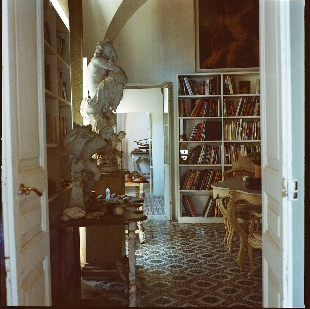 manfredi gioacchini's photo along the open enfilade doorways of cy twombly's house in gaeta, where the library dominates between two sets of doors. along the left wall are tables and a pedestal with white classical sculptures on them, surrounded by crystals, shells, and other tchotchkes. beyond the library through the farther doors is what looks to be table with hats on it, and another door beyond that. the sculpture closest to the foreground was photographed head-on by tacita dean in 2008, and is the subject of this whole effort. though it is also interesting to look through all the photos gioacchini made for other details.