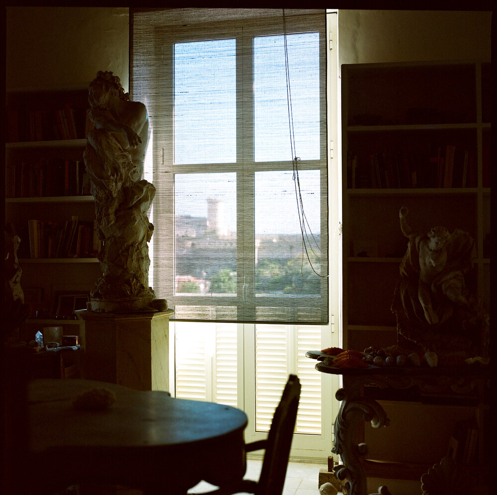 a dark interior and bright sunny window view in manfredi giaocchini's photo of cy twombly's library in gaeta. in the shadow, two white classical style sculptures stand, the arger one on a pedestal, on the left, and the shorter one, on a tchotchke-filled table, on the right. a dark tabletop in the foreground reflects a little sunlight, but is out of focus