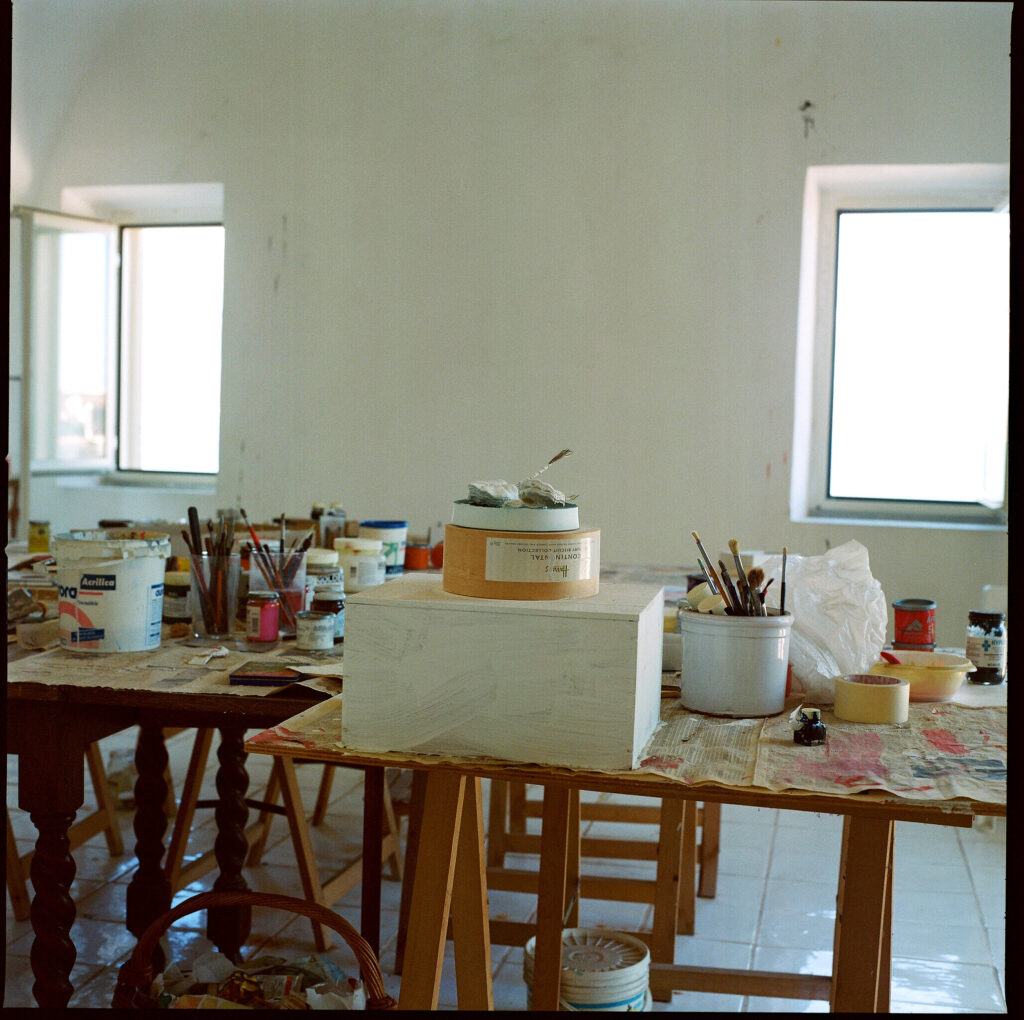 a photo of cy twombly's painting studio in gaeta with supplies as he left them on rickety tables, and an unfinished sculpture-like tower of boxes in the center. the view of the bay out the windows is washed out by the exposure. manfredi gioacchini fecit.