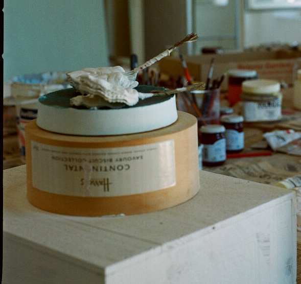 a detail from manfredi gioacchini's photo showing the unfinished sculpture in cy twombly's studio, a yellow round box of harrods savory biscuits, upside down on a white painted wood box. a green-topped pie plate looking thing is on that, wht these glove and stick, or rib and pouch or who knows what fetishy little objects are perched on top