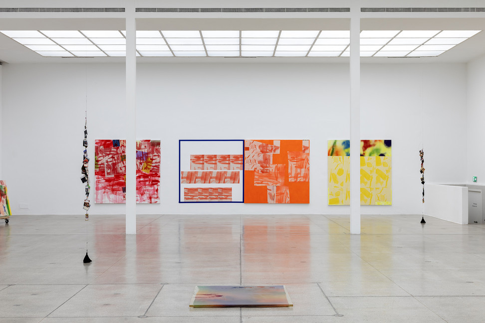 a concrete-colored granite slab floored gallery with a bright white grid of overhead lighting and a stairway in the corner at the secession in vienna is installed with several large square paintings in red, orange, and yellow, on the far wall, plus a square of the same size marked out in blue tape containing three rows of smaller images. two strings of laminated postcard-size photos hang from the ceiling; a cart loaded with some rainbow palette abstract paintings is in the left corner, and an indistinct object in a swirl of color and solid acrylic slab is on the floor in the lower foreground, an installation view of rochelle feinstein's 2024-25 show, the today show