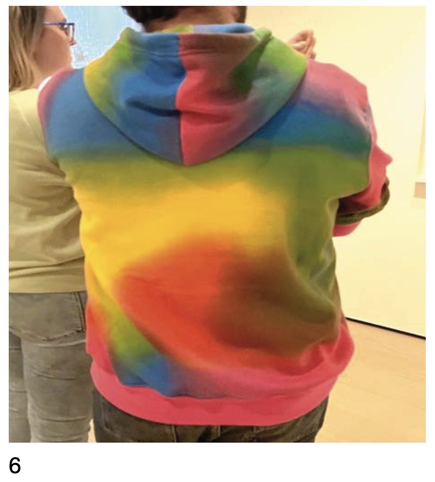 a cropped photo taken from behind some haples white guy with dark hair, who was just minding his own business while  wearing a swirly rainbow gradient hoodie, and a white lady with no rainbow clothing standing to his left, at moma in 2023, when rochelle feinstein had just about enough rainbows, and snapped this photo.