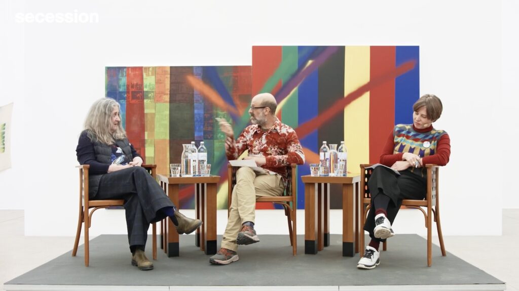 three white ppl sitting on a very low dais in viennese chairs with viennese side tables between them: from left, rochelle feinstein, in dark clothes with long, curly hair parted in the middle; in the middle justin lieberman, a bald bearded thin fellow in khakis and a festive red printed shirt, is pointing toward feinstein; on the right, stephanie weber, younger-seeming, with short bobbed brown hair, also parted in the middle, black pants and a geometric sweater. all have their legs crossed. behind them is a two-part painting by feinstein whose main element is vertical stripes of primary/rainbow/or tv test pattern colors, crossed multiple times with thick passes of sprayed color of the same palette. it is a screenshot of a conversation from december 2024 at the secession in vienna.