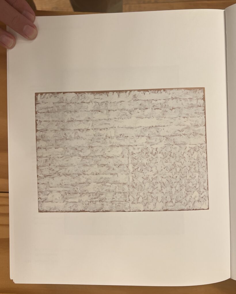 a photo of a white painting on paper by jasper johns of a us flag, p32 in the catalogue raisonne, a 1957 work in the collection of the metropolitan museum, turned upside down to become a study for an upside down repetition of a johns flag painting.