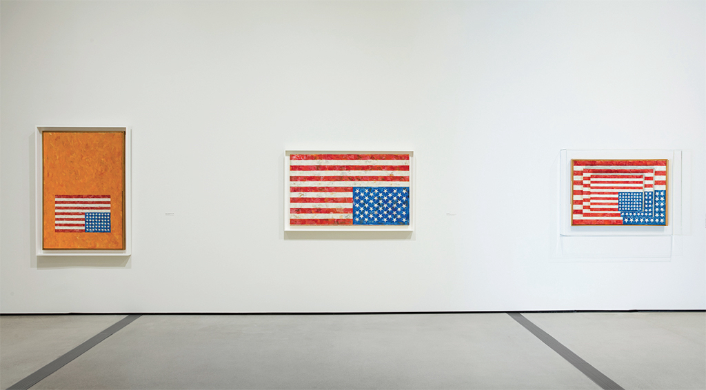 an installation view of three jasper johns flag paintings on a white wall with a dark floor, from a 2018 show at the broad collection in los angeles, but each painting has been turned upside down. original image by eugenio rodriquez via artforum