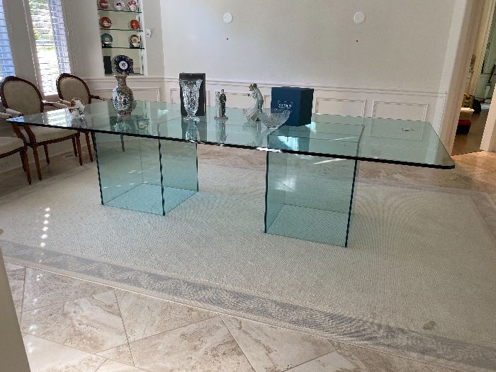 a glass dining table with a wavy edge sits on two square glass bases sits on a beige sisal area rug in a beige travertine floored beige dining room where the sconces have been removed from the beige wall above the beige wainscotting. supposedly this table was used in filming the final scene of john hughes' 1984 date rape teen comedy Sixteen Candles, where jake the rich senior, after trading away his passed out girlfriend to be raped by anthony michael hall's geek, sits on this table with molly ringwald's character, a sixteenth birthday cake in between them. the estate sale manager of this highland park illinois sale claimed without any documentation that this table moved from the movie location house to its c.2020 house, though nothing else in the estate seems to indicate any connection to hughes or the film.