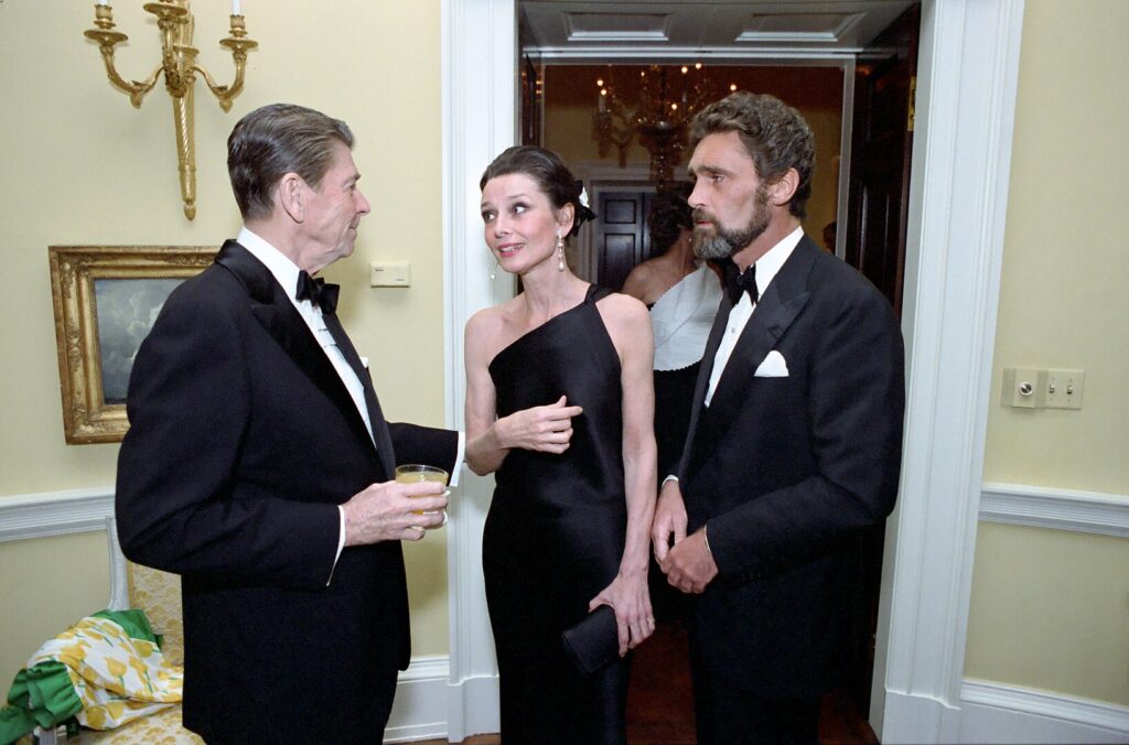 ronald reagan on the left, audrey hepburn in the middle, and robert wolders, her boytoy on the right, all in black formalwear, in a doorway in the white house, a room yellow walls which has probably been spray painted gold by now, don't get me started, in a 1981 white house photo. someone's yellow printed silk evening wrap with green ruffled edges is draped over the arm of the white painted french style chair behind reagan's ass.