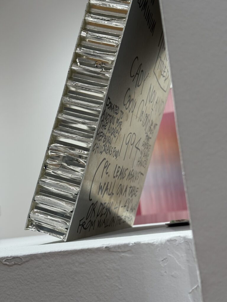 a closeup photo of the side and back of a cady noland sculpture on view at christie's in may 2025. the work is a laptop-sized panel of honeycomb filled aluminum resting at an angle on a white plinth against a white wall. on the back is written in black sharpie: the artist's name and signature, a box that says not titled,' a date, 1994, donated to the audrey hepburn children's' fund, a warning not to touch the front surface with bare hands, and detailed instructions of how to install it: please lean against wall on a table or a desk, 4 1/2 - 4 3/4 inches from the wall at the bottom, photo via ian ware