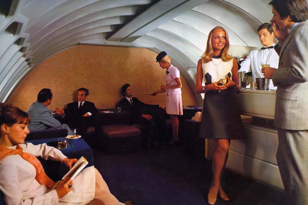 a 1970 air france brochure image of the upper deck lounge on the new 747, in which a white couple stand smoking and drinking in front of a white jacketed (and white) bartender on the right, a white lady with an orange scarf sits reading on the left foreground, and the back wall of the lounge is a cream colored silk bas relief panel by sheila hicks, which extends the full width of the curved and ribbed cabin ceiling and walls. three white guys are lounging while a pink clad blonde flight attendant takes their order. the point of all this at this moment is to document the hicks panel in situ. she made 18, or 19, and delivered 18, and kept one, i think