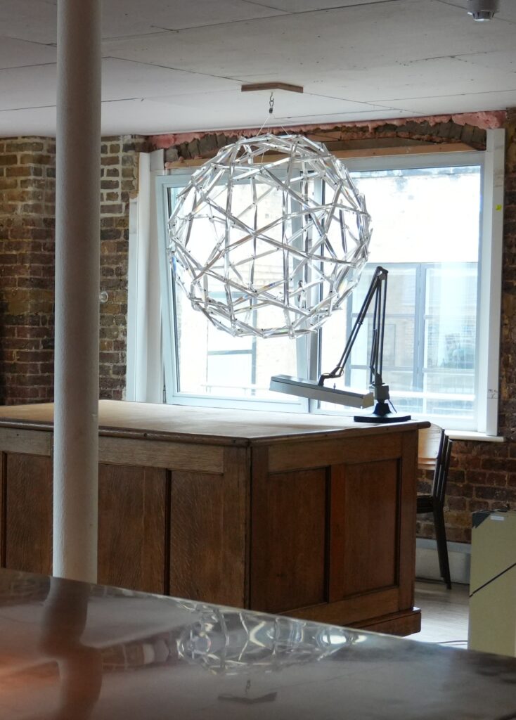 a lattice of long, thin, acrylic prisms held together at the ends by fishing wire froms a truncated icosahedron sphere, which buckminster fuller called a chandelier. almost a meter in diameter, it hangs over a deep brown desk in an artfully rough brick space, reflecting light from the wide window behind it. a draftsman's articulated lamp is clamped to the edge of the table. the chandelier is reflected in a polished tabletop in the foreground, and a single column runs along the left side of the photo, which was cropped from a larger image taken in the drawing matter archive in central london by jesper authen. 