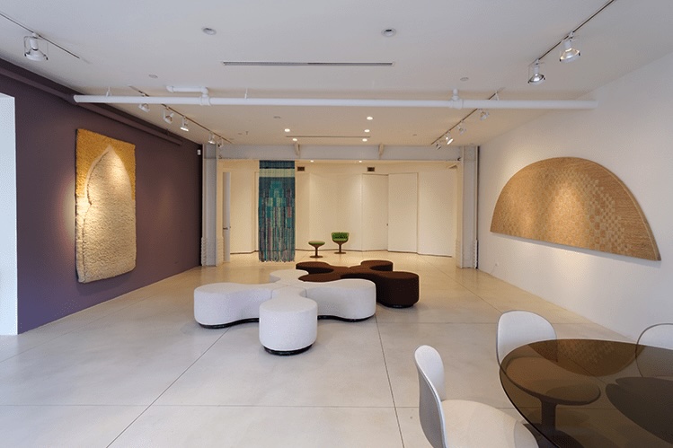 two cream to tan textile works by sheila hicks hang on walls facing each other in a marble tiled gallery space. in the center are interlocking settees in the shape of a four pointed star with bulbous tips. the shape is like a chew toy, or the cingular logo, but neither reference feels especially helpful here. a smoky glass table with white mod chairs sits in the lower right corner of the image, and a hicks piece of cascading turquoise hangs from the ceiling at the far back. a 2014 exhibition at nyc furniture/design gallery demisch danant