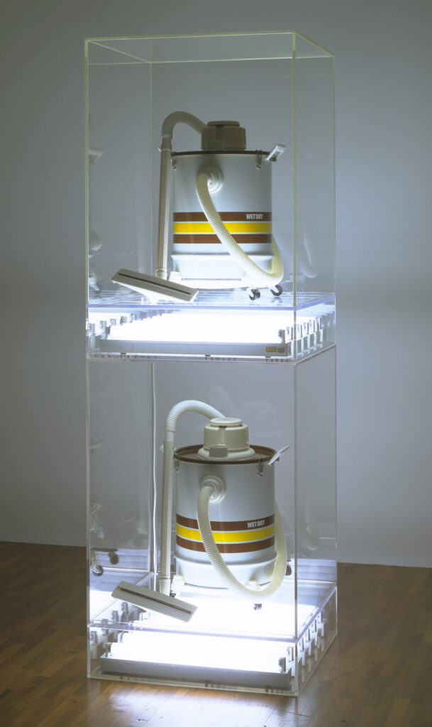 a jeff koons sculpture of two wet/dry canister vacuum cleaners, white with red and yellow stripes around their middle, inside a two-level plexiglass case. a row of white fluorescent lights blazes beneath each vacuum cleaner. from moma's collection.