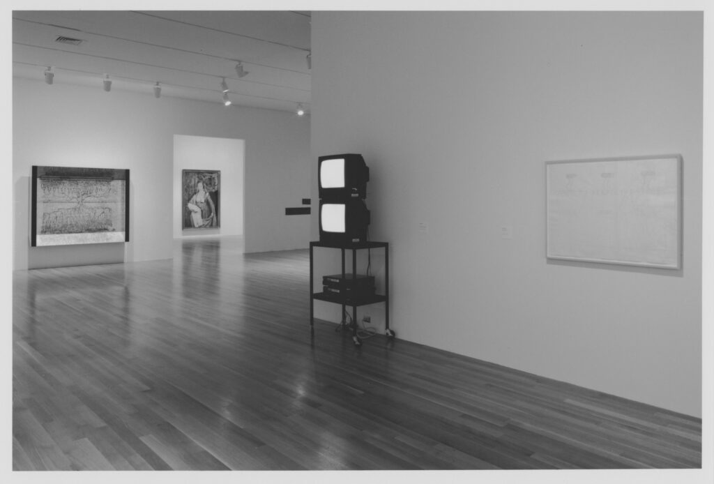 a black and white installation photo of a 1997 show at moma is mostly empty white cube gallery space and a gleaming hardwood floor. an illegible drawing hangs on the right wall, an illegible drawing in some kind of shadow box hangs on the wall in the middle background, and in the distance through a doorway, an illegible cindy sherman photo of herself as a nun or saint or whatever, so all that can really be made sense of here is the bruce nauman work at the center of the image: two video monitors stacked on top of a black rolling cart, and two laserdisc players stacked on the shelf beneath. the monitors are glowing, but the images are illegible. i swear, this show was more interesting in person.