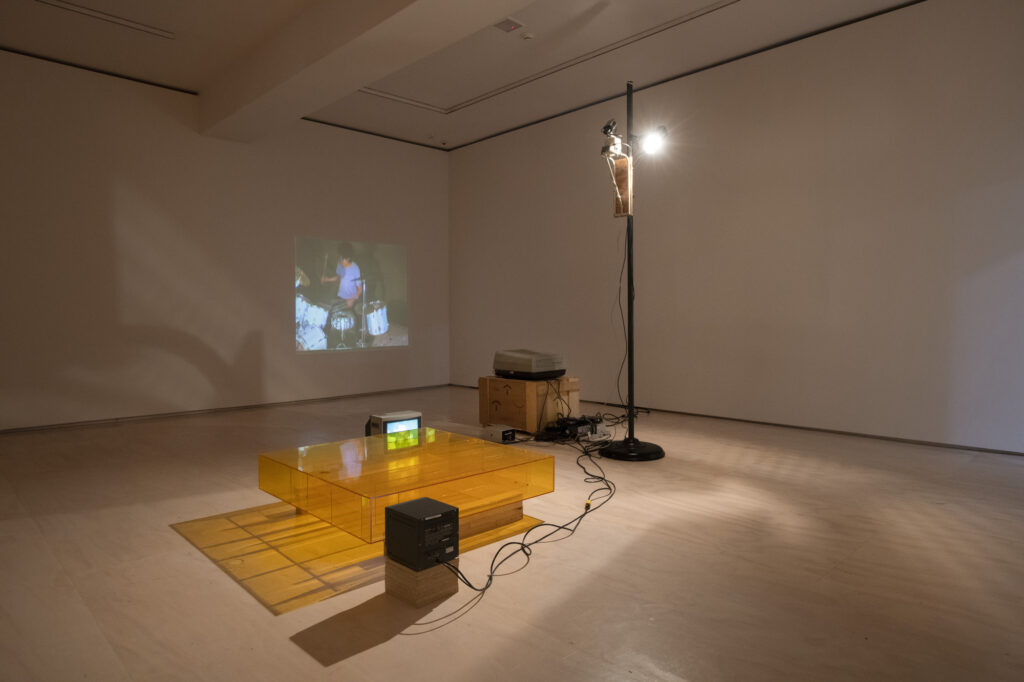 in a dimly lit white cube gallery, two video monitors are set with their screens facing either end of an enclosed rat maze made of piss yellow plexiglass. video cameras and a spotlight on a tall stand are pointed down onto the empty maze. a 1980s video projector on a wooden pedestal projects interspersed images of a white teenage boy wailing at a drumset, making an incredible racket; closeup shots of a rat running through the maze; and live images of the now-empty maze, and sometimes viewers' feet as they enter the frame. by bruce nauman, in moma's collection since 1996.