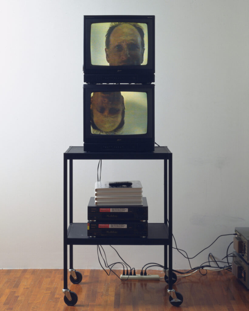 two square video monitors in black cases are stacked on a thin black metal rolling cart. on the shelf below two black panasonic laserdisc players fresh from the box are stacked on each other, with four white vacuum molded plastic clamshell folders for holding laserdiscs are on top of the players. a single black remote control sits atop that. a tangle of black cables connects to a generic powerstrip on the maple strip floor of a soho loft, and a couple of cables snake off to the right edge of the image, perhaps to a transformer. on the monitors, the tightly shot head of a middle aged white guy, the artist bruce nauman is captured in a single frame. the head on the lower monitor is upside down. this 1993 work, think, by bruce nauman, entered moma's collection in 1996, but this image has to predate that.