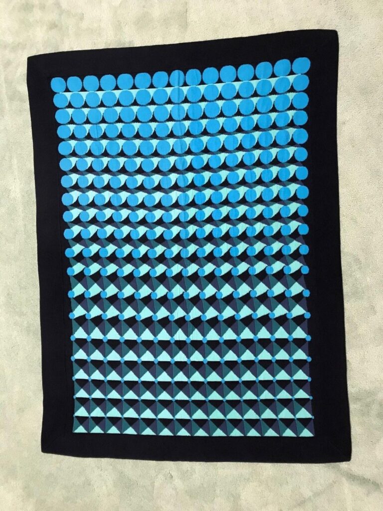 a black-background machine knitted wool blanket with a smooth black border has a geometric progression pattern of green squares and triangles developing into aqua circles. this lap blanket was designed by olafur eliasson for netjets europe customers in 2005, and someone flipped this one on ebay at some point, which led to its being archived on worthpoint, photographed on the floor for sale