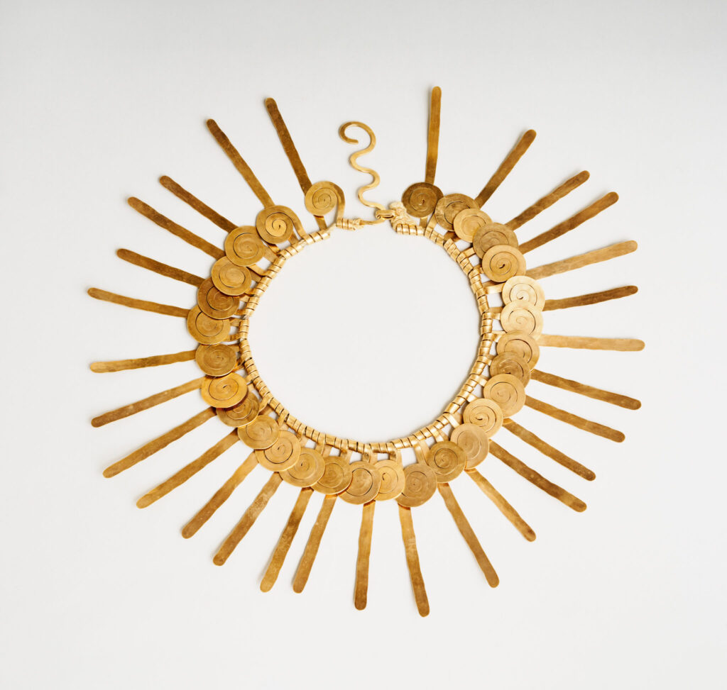 an alexander calder necklace of flattened gold wire from 1937 is laid out flat in a circle. strips of gold wire twisted around a central cord are alternately turned into flattened spirals, or left straight, so they radiate outward. laid flat like this, it looks like a child's drawing of the sun. from the calder foundation