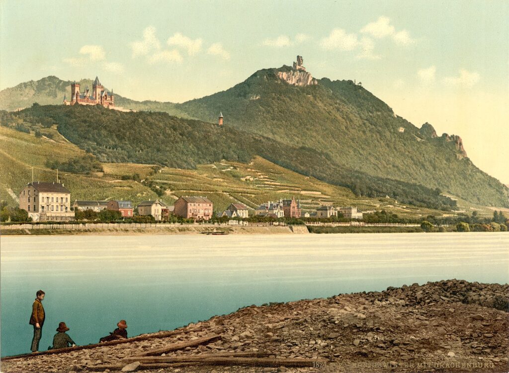 a 1900 postcard with weirdly intense colors, depicting the upper rhine valley site of drachenfels, a mountain over the river with a medieval ruin on top. on a lower hill next to drachenfels is a c.1883 neo-gothic castle built by some prussian aristo, and below that, on the river's edge, are the buildings of the town of koenigswinter. at the very bottom of the image, on the opposite bank, are three figures, the whole thing feels made up, which, well, yes and no