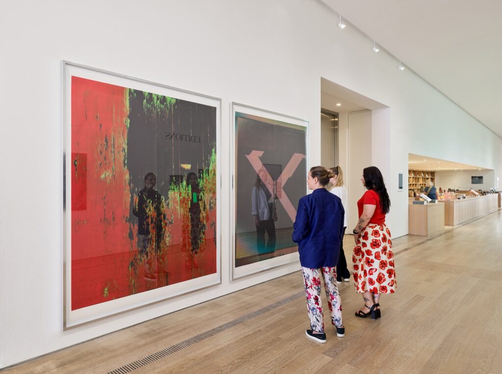 three white women in swiss festive clothing stand in front of two wade guyton prints that reproduce two paintings at full scale. the abstract one on the left in red and black uses a photo of the artist's studio floor; the one on the right is a scan of an x painting. both have been digitally manipulated. they are in thin steel frames in the lobby space of a museum, the fondation beyeler, in basel in 2025