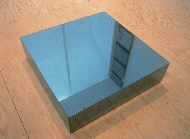 a square sculpture eight inches high made of blue mirror sits on a plywood floor, reflecting a skylight and bookshelf in the lehman maupin gallery in this 1997 photo of felix gonzalez-torres' 1992 work "Untitled" (Fear).