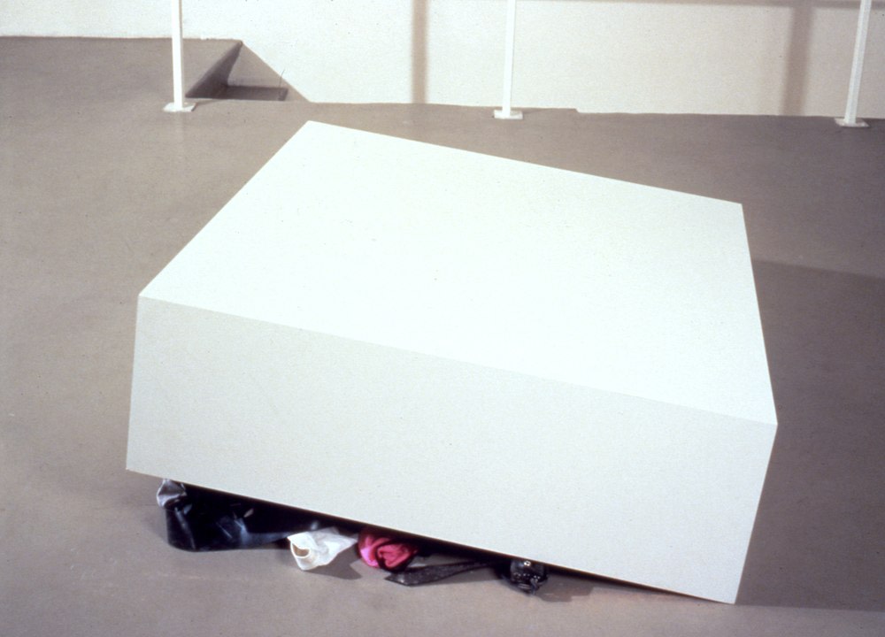 a slightly off white painted wood pedestal a foot high and 40 inches on each side has one side propped up like a box trap, with underwear and some harnesses peeking out from below, a non-work by felix gonzalez-torres photographed ni the mezzanine at galerie ghislaine hussenot in paris in 1992