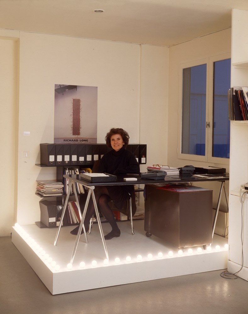an elegant white lady sits at a chrome and black desk with office accoutrements around and behind her, on a wooden platform that fits into the corner of her art gallery office. the pedestal is painted pale blue and has white light bulbs along the two outer edges. a non-work by felix gonzalez torres installed in 1992 at galerie ghislaine hussenot in paris