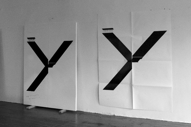 two mostly identical black and white artworks photographed in black and white against a white wall: a painting of a giant, misprinted capital x on a stretched canvas sitting on two little blocks is on the left, and the fundraising poster version of it, same size but with creases from being handfolded, and on paper, is on the right. the last of six fundraising edition posters by wade guyton for printed matter, 2019