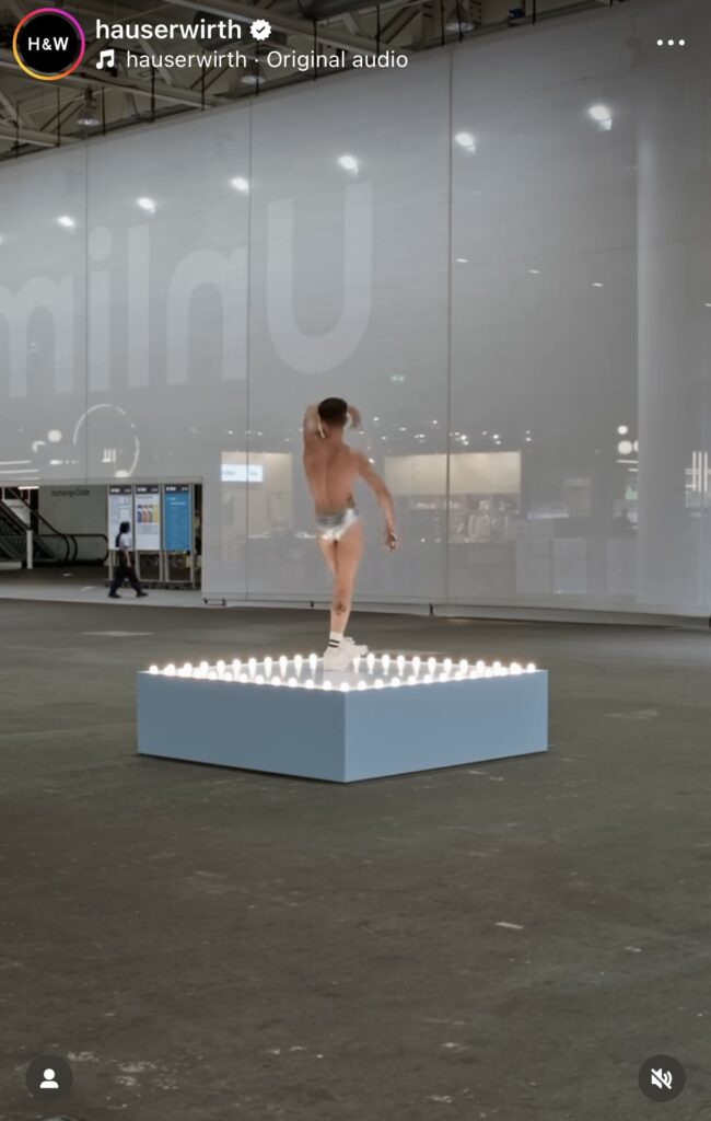 a screenshot of hauser & wirth's instagram of a video of a white go-go dancer in a very small silver speedo and sneakers, facing away from the camera, dancing to himself on a pale blue square platform ringed with soft white light bulbs, installed in a vast concrete convention center, with a scrim in the background, the messe, for art basel. a work by felix gonzalez-torres shown by a gallery that does not officially represent the artist
