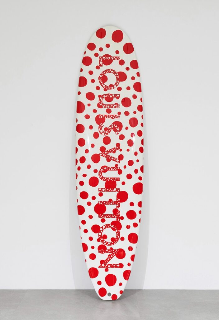 a white surfboard with red dots all over it and the name louis vuitton running down the center in tighter red dots, leans against a white wall. being sold at sbi auction in japan in july 2025
