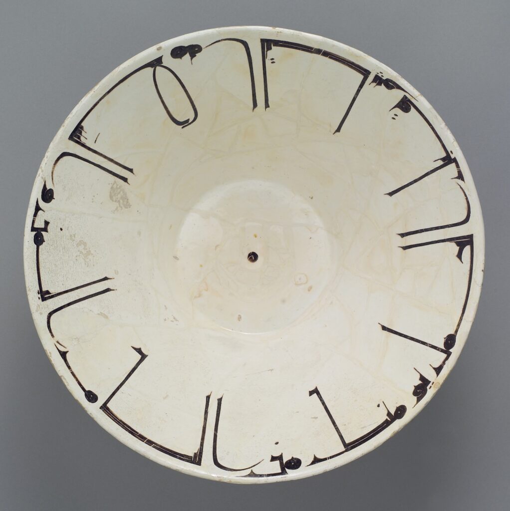 a top down view of an 18 inch diameter white porcelain bowl with thin, geometric, elongated, almost abstract kufic-style arabic calligraphy around the rim, with extensions down into the bowl, written in black slip. from 10th century iran, at the met