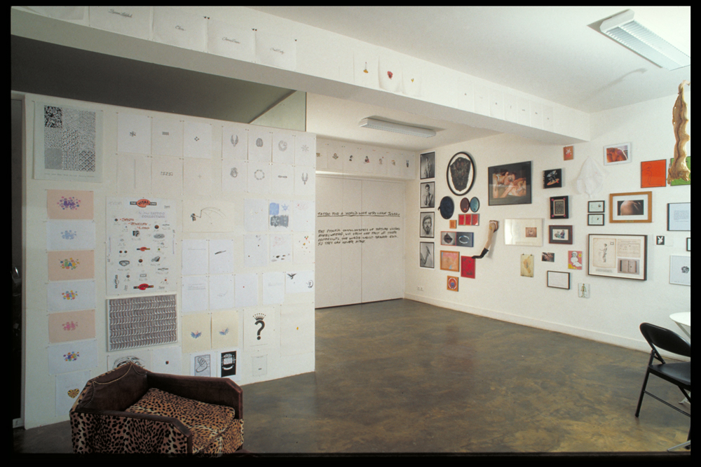 a low ceilinged art gallery with white walls and a concrete floor is filled with small works on paper, mostly tattoo designs created by artists. a june 1992 exhibit at air de paris gallery 