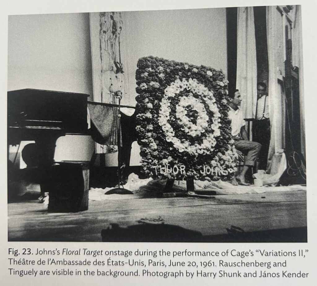 a 1961 shunk & kender photo of a 1m or larger square flower arrangement in the form a jasper johns target sits on a stage in paris, a grand piano in the rear left, and robert rauschenberg and jean tinguely in between curtains and backdrops on the right, from jasper johns's catalogue raisonne, vol. 5