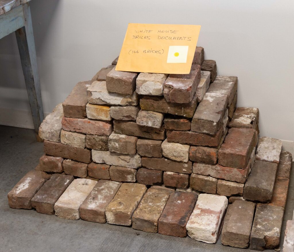a pile of 18th and early 19th century bricks in a variety of finishes sits in a five-layer stack on the floor of a rural virginia auction house. a manila envelope on top of them has white house brick documents 166 bricks written on it. because these bricks are from the white house.