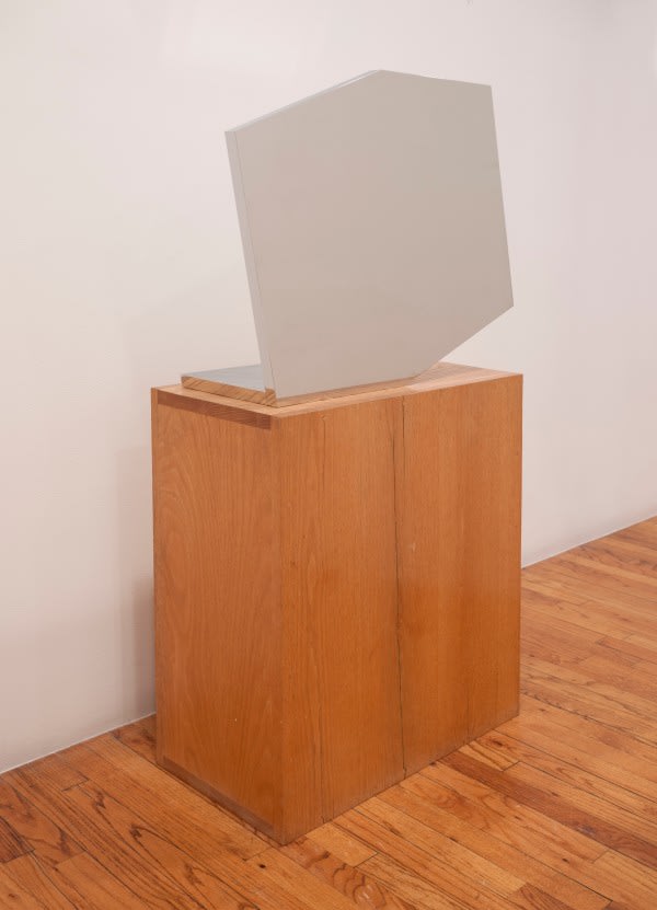 brooke alexander's photo of ellsworth kelly's mirror finish stainless steel sculpture in the shape of a truncated rectangle sits on a maple stained wood pedestal in a gallery loft space. the angle of the photo is such that the sculpture's mirror surface is entirely neutralized, and seems monochrome grey. via art basel