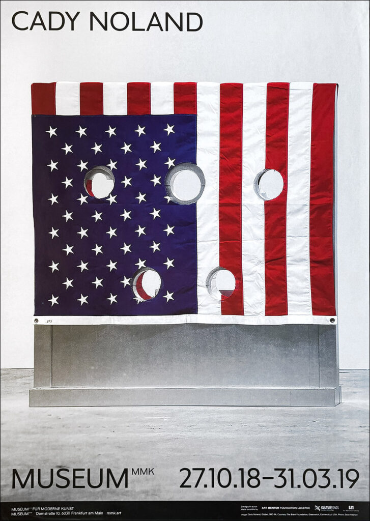 a poster for cady noland's 2018-19 exhibition at mmk frankfurt has the artist's name and date over a full bleed photo of gibbett, a 1993-94 work that is an aluminum stockade for a single incarcertant, with five holes for their head hands and feet, and an american flag draped over the stockade, with holds cut to match on both sides. the side facing the camera is the canton of stars. this example of the poster is for sale at bent priorities dot com as of 2025