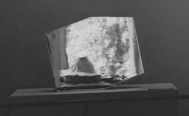 a detail of james mathew's 1971 installation photo of moma's exhibition, techics & creativity II, showing ellsworth kelly's truncated rectangle-shaped mirror-finish plated steel sculpture, Mirrored Concorde, reflecting a volcano-looking claes oldenburg sculpture of a respiring ice bag, in front of the large plate glass windows by which the gallery connects to the sculpture garden