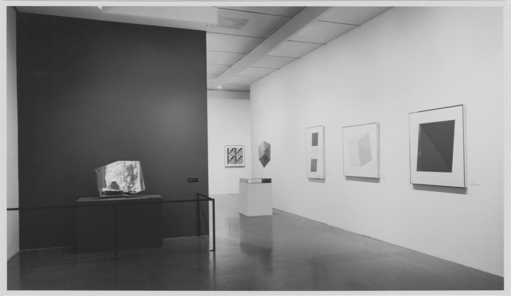 james mathews' 1971 installation photo of a moma print exhibition is mostly the work of ellsworth kelly. a series of kelly prints is mounted on the white wall along the right, and kelly's mirrored concorde sculpture is on a pedestal, behind a stanchion, and against a darkly painted accent wall on the left.