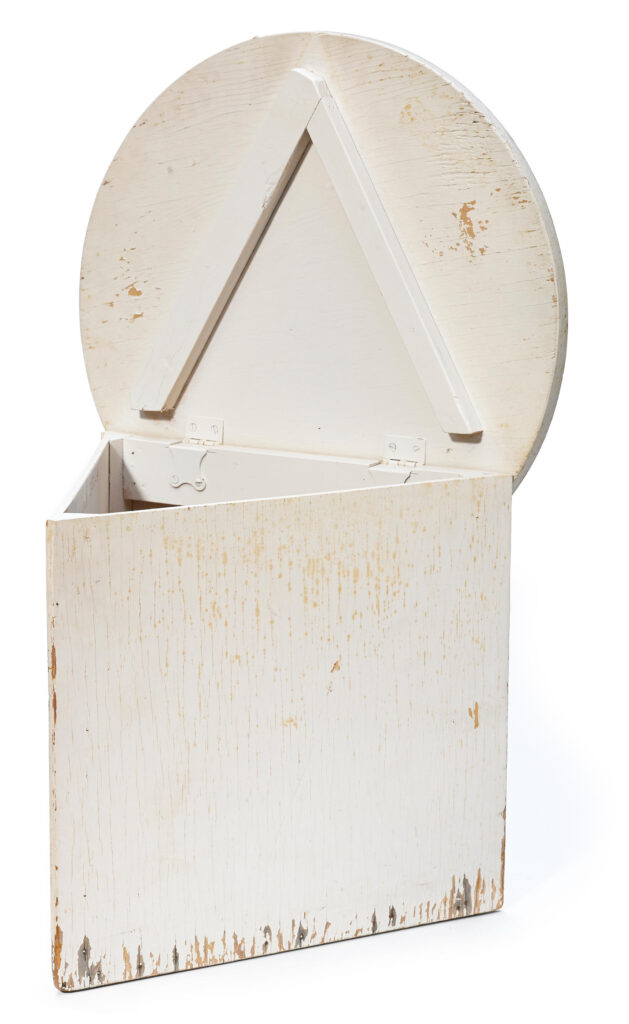a triangular box table with its circular lid open and hinges visible along one side, all painted long ago in white that's scuffed, chipping, and leaching while the 90-yr old plywood underneath starts to splinter too. this image is from 2014, when bonhams sold this original rudolph schindler table for $6000.